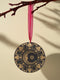 Round metal ornament featuring a golden crescent moon face with star detail, surrounded by art deco shell motifs on black background. 3.5-inch diameter double-sided design with lunar phase pattern and crimson ribbon attachment. Vintage-inspired celestial artwork.